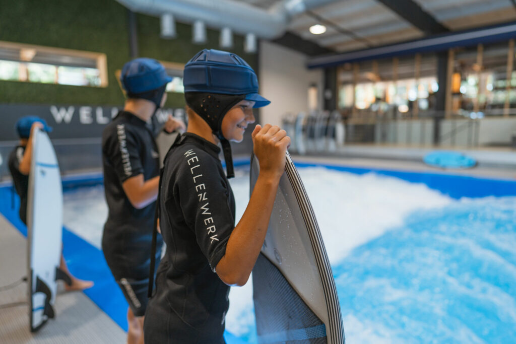 Glücklicher Junge mit Surfbrett und Helm steht am Rand der Indoor Welle und schaut ins Wasser